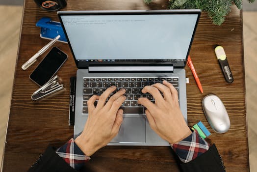 Overhead view of hands typing on laptop surrounded by office supplies on a wooden desk.